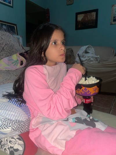 A young female child is depicted seated indoors in a domestic setting within Anápolis, Brazil. The child, possessing long dark hair, is attired in pink long-sleeved pajamas featuring a patterned lower section with geometric and character designs. Her gaze is directed towards the right side of the frame, with her mouth slightly open, indicating focus. She holds a black, Halloween-themed popcorn bucket, adorned with purple and orange haunted house and bare tree motifs, filled with popcorn. This bucket is functionally combined with a Coca-Cola branded bottle/cup, into which a black straw is inserted. The child is positioned on a surface covered by a white and blue striped blanket and a red blanket section displaying a white Mickey Mouse head silhouette pattern. To her left, a patterned pillow or cushion is visible. The background consists of light blue walls. A framed image depicting a dark subject is mounted on the left wall. On the right wall, another framed picture, showing a landscape, is present, with a partial view of a wall clock above it. A light-colored sofa, draped with a crumpled light-colored fabric, is situated against the right wall. The ambient lighting is dim.