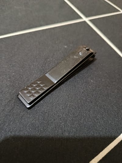 A dark-colored nail clipper is resting on a black textured surface, which appears to be a fabric with a grid pattern. The nail clipper is positioned diagonally across the frame, with its textured grip facing the bottom left. The surface is illuminated from the top left, casting a subtle shadow to the bottom right. The overall scene is uncluttered and focuses solely on the nail clipper. There is no indication of the specific location within Depok, Indonesia, or any people or actions taking place.