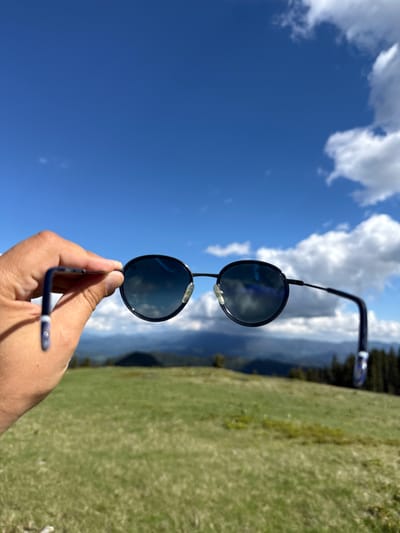 A hand holds a pair of dark-rimmed sunglasses up to the sky, obscuring the view of a mountainous landscape below. The sunglasses have round, dark lenses that reflect the blue sky and white clouds. The hand is in focus, with visible skin texture and fingernails. The background is a panorama of rolling green hills and distant, dark mountains, dotted with evergreen trees. The sky is a vibrant blue, with scattered fluffy white clouds. The image appears to have been taken outdoors in a natural setting, possibly in Gela, Bulgaria, a village known for its scenic mountain views.