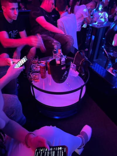 The image depicts a group of young adults at a table in a dimly lit venue, possibly a nightclub or bar, with purple and blue lighting. In the foreground, a person's hand is visible holding a smartphone displaying a screen with app icons. On the round table, there are several drinks, including two glasses with amber liquid, a can of Coca-Cola, a bottle of Johnnie Walker, and an ice bucket. Ashtrays are also present on the table. Several individuals are seated or leaning around the table. The overall atmosphere appears to be social and relaxed. The image does not provide any specific clues to suggest it is located in Gabrovo, Bulgaria.