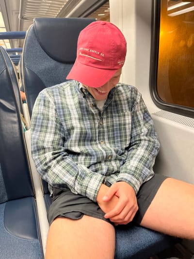 The image depicts an individual seated on a public transit vehicle, likely a train, characterized by blue upholstered seating and white interior paneling. The individual, presumed male, wears a red baseball cap with "JETTY SURF SUPPLY CO." and a surfboard graphic, obscuring his eyes and forehead; his mouth is visible, opened in a smile. He wears a long-sleeved plaid shirt in shades of blue, green, and white over a lighter undershirt, and dark shorts. His hands are clasped on his lap, and a dark band is visible on his left wrist. To the right, a window reveals an indistinct orange-brown illumination or reflection from outside, with a metallic grille below. The specific location within Red Bank, United States, cannot be determined from the visual evidence provided.