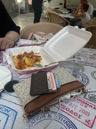 In Budapest, Hungary, at an outdoor cafe, a table is set with a takeaway food container, a plastic fork, a small glass jar, sunglasses, a wallet, and a purse. The food container holds fried pieces of food in a sauce. The wallet contains several cards, including an identity card from the Republic of Bulgaria. The purse, a brown leather and beige Michael Kors design, has charging cables protruding from it. The table is covered with a cloth featuring passport stamps. In the background, two individuals are seated at another table, and a person is standing. The overall setting suggests a casual dining experience.