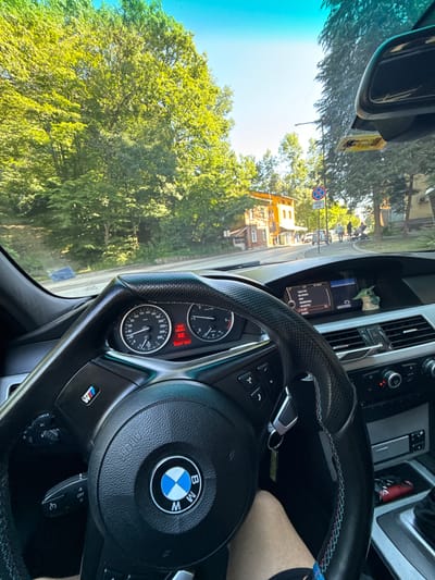 The image captures the interior of a BMW vehicle, viewed from the driver's perspective, as it navigates through the city of Gabrovo, Bulgaria. The steering wheel, a prominent feature, displays the BMW logo and M-sport badging, indicating a performance-oriented model. The dashboard shows the car's instruments, with the speedometer indicating a speed of approximately 60 km/h and the digital display showing the time as 9:27 and a temperature of 22.5°C.

Outside the car, the scene is characterized by lush green trees lining the street, suggesting a pleasant, sunlit day. The road ahead is paved, with a sidewalk and buildings visible on the right side. One building, with an orange facade, appears to be a shop or residential property. Several pedestrians can be seen walking on the sidewalk and crossing the street in the distance, alongside a few parked and moving vehicles. A blue and red circular traffic sign is also visible on a pole. The sky is a clear blue, further enhancing the daytime setting. The car's interior also features a small figurine of "The Child" (Grogu) from "The Mandalorian" placed on the dashboard.