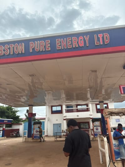 This image depicts a street view of a gas station in Obosi, Nigeria, under an overcast sky. The primary subjects are two individuals, one wearing a blue shirt and denim shorts, and another in a black garment. They are near gas pumps. The gas station has a prominent sign that reads "BSTON PURE ENERGY LTD" in bold, red lettering against a blue background with red trim. Another visible sign reads "Sabston" on a gas pump.
The scene is a typical street-level view of a commercial establishment. The architecture suggests a building with multiple windows and doors. Reflections on the shiny surface of the gas station roof indicate a diffused light, consistent with an overcast day. The ground is paved, and a motorcycle is parked to the side, suggesting a bustling environment where people use personal transportation. The overall impression is that of a functional, everyday setting in a Nigerian town. The people's postures suggest casual engagement, possibly refueling vehicles or interacting at the station. No specific time of day is strongly indicated beyond the general daylight.