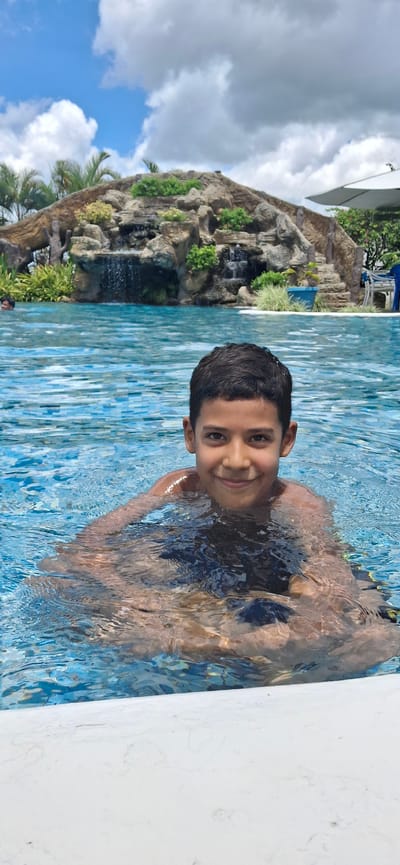 In this media file, a young boy is pictured in a swimming pool, with a lush, rocky waterfall and greenery in the background. The boy, with dark hair and a warm smile, is partially submerged in the blue, rippling water, his shoulders and upper body visible. The sunlight casts reflections on the water's surface, creating a dynamic texture.
Behind him, a man-made waterfall cascades down a rocky formation, adorned with vibrant green plants. Palm fronds are visible, suggesting a tropical or subtropical climate. The sky above is a mix of bright blue and fluffy white clouds, indicating fair weather. The scene evokes a sense of relaxation and enjoyment, likely during a warm, sunny day.
The overall impression is one of a fun recreational activity in a visually appealing, resort-like setting. The image appears to be taken outdoors, with natural light illuminating the scene. No visible text is present in the media file.