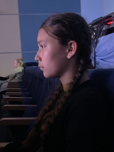 A close-up, profile view shows a young girl with long, braided brown hair sitting in a dark theater. Her head is turned to the left, and she appears to be looking at something off-camera. The background is blurred, but it is possible to make out rows of blue seats and a wall with blue and white tiling. Another person, a young woman with blonde hair pulled into a bun, is visible in the background, seated in one of the rows. The lighting is dim, with a pinkish hue cast on the girl's face. The image is likely taken in a movie theater or auditorium in the city of Donskoye, Russia.