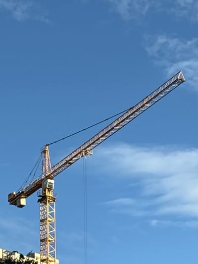An image shows a yellow tower crane against a clear blue sky with some white clouds. The crane is positioned in the city of Pushkino, Russia, and is a prominent feature of the construction site. The crane's jib extends diagonally across the frame, with a counterweight visible on the left side. The main tower of the crane is vertical, and the operator's cabin is situated near the top of the tower. The lower part of a building is visible in the bottom left corner of the image, partially obscured by trees. The scene is static, with no people or ongoing activities depicted.