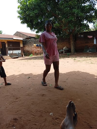 The image shows an outdoor scene in Ogidi, Nigeria. A woman wearing a pink T-shirt and striped shorts is walking across a dirt yard toward the camera. She is wearing flip-flops. To the left, a young boy wearing shorts and flip-flops is partially visible. In the foreground, a pair of feet are visible. In the background, there are buildings constructed of brick and corrugated metal roofing. A tree provides shade. Clothes are hanging on a line in the background. The ground is dirt, and there is debris scattered across it.