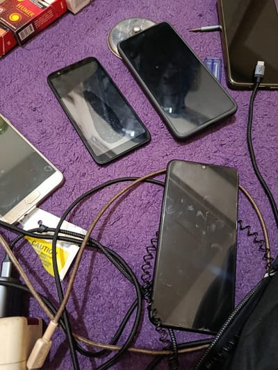 A still life photograph depicts several electronic devices, including three smartphones, charging cables, and a power adapter, arranged on a textured purple surface that appears to be a rug or blanket. One smartphone, a black model, lies face down with its screen scratched and smudged. Another, a gold-colored device, is positioned horizontally with its screen facing up and also exhibits smudges. A third black smartphone, also with a smudged screen, is oriented vertically. Several charging cables, one with a "120W" marking, are intertwined with the phones. In the background, a pack of "HUMER" brand cigarettes and a small, circular metal object are visible. The overall impression is one of personal effects, possibly in a domestic setting. The image is taken from a high-angle perspective. The specific location of Depok, Indonesia, cannot be determined from the visual information provided.