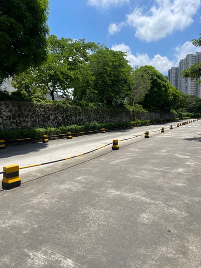 A sunny day with a blue sky and scattered clouds, the image shows a paved road lined with yellow and black traffic delineators. A stone wall with lush green foliage growing on top borders the road on the left. Beyond the wall, trees are visible. In the background, modern high-rise buildings stand tall. The road appears to be a pathway or lane, possibly within a residential or commercial area in Panyu District, China. There are no people or vehicles present in the scene.