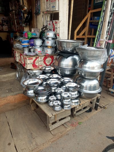The image shows a street-level view of a small shop in Arawa, Nigeria, specializing in metal kitchenware. In the foreground, a wooden stall displays stacks of various sizes of shiny, silver-colored metal bowls and pots. Some of the bowls are nested within each other, while others are stacked on top of boxes. The boxes, branded with "TOSIBA BRITE ALUMINIUM COOKWARE," are visible beneath some of the displayed items. Behind the stall, a selection of colorful plastic containers and more metal cookware can be seen. The shop appears to be open-air, with a rough, reddish-brown textured wall on the left and a wooden structure and bundled materials on the right. The ground is a mix of worn wooden planks and rough concrete or dirt. The overall impression is one of a bustling marketplace stall.