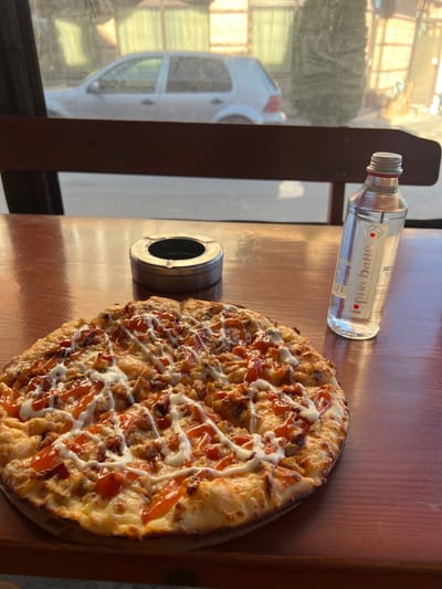 On a wooden surface, likely a table, are arranged a pizza, a small glass bottle, and a metal ashtray. The pizza is round, topped with what appears to be meat or vegetables, and drizzled with white and orange sauces. The glass bottle is small, possibly containing a liquid, and features a label in a non-English script. The metal ashtray is cylindrical. Outside a window, a glimpse of a white car and greenery suggests an exterior setting. The scene is illuminated by daylight. The information about Breznitsa, Bulgaria is not evident in the image.