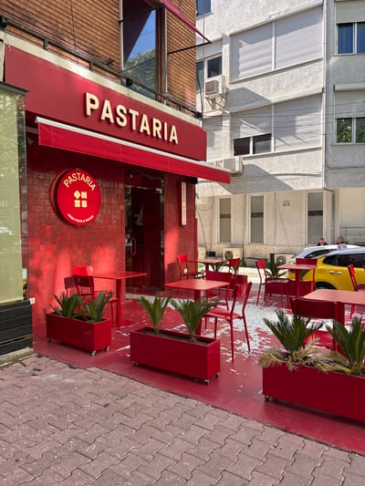 The image shows the exterior of "Pastaria," a fresh pasta restaurant located in Tirana, Albania. The facade is primarily red, with red tiles, a red awning, and a red sign featuring the restaurant's name in white lettering. A round red sign also displays the name "Pastaria" along with the words "Fresh Pasta & Sauces." Red tables and chairs are arranged on the red tiled area in front of the restaurant, interspersed with red planters containing small palm-like plants.
The buildings surrounding the restaurant are primarily white, and some have windows with closed blinds. A yellow taxi is visible parked near the outdoor seating area. Pedestrians can be seen in the background near the taxi. The image is taken during daylight. The sidewalk adjacent to the restaurant is paved with interlocking brick tiles.