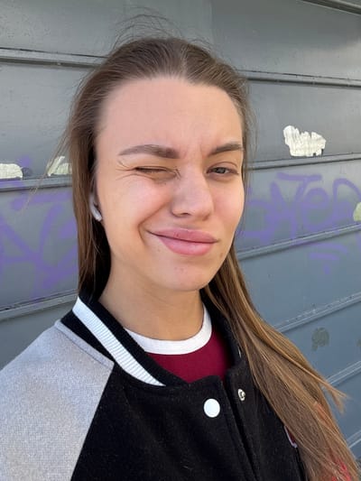 A young adult woman is shown in a portrait-style photograph. She is winking with her left eye and smiling. She has light brown hair and is wearing a maroon shirt underneath a black jacket with white and black striped trim. She appears to be wearing a white wireless earbud in her left ear. The background shows a gray metal surface with some purple graffiti and a sticker. The location appears to be an urban area, potentially Liepaja, Latvia, but there are no specific elements to confirm this city.