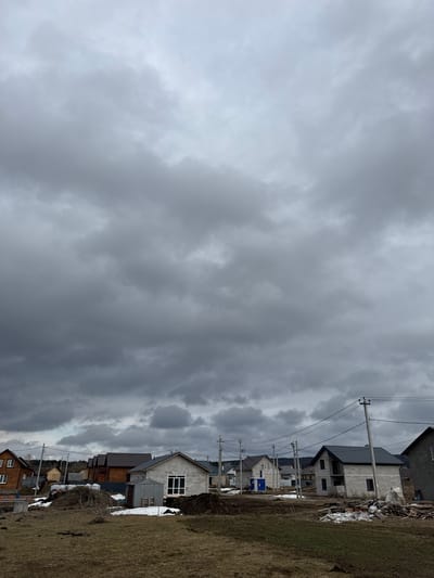 The image depicts a cloudy landscape in Gornyi, Dvurechenskoe S/P, Russia. Several houses are visible on the right, varying in construction and materials. Some appear to be newly built with unfinished exteriors of gray concrete block, while others are older with wooden facades. Power lines and poles run parallel to the row of houses. The ground is covered with dry, brown grass and patches of snow. The sky is overcast with heavy, dark clouds. No people are visible.