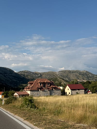 The image captures a rural scene in Kopito, Montenegro, under a bright blue sky with scattered white clouds. In the foreground, a dry, tall grass field stretches out, bordered on the left by a paved road with a white line marking its edge. A small white marker with a red top stands near the roadside.
Dominating the middle ground are several buildings. The largest is a substantial house with a multi-tiered, reddish-brown tiled roof and stone-clad lower walls. It has several windows visible. To its right, a smaller, simpler building with a red roof and pale yellow walls is situated. Further to the right and behind this, a tiny red-roofed structure is visible. Green trees and shrubs dot the landscape around the buildings.
In the background, rugged, rocky hills rise up, covered in sparse vegetation. The terrain suggests a mountainous or hilly region. The lighting indicates it is likely daytime, possibly late afternoon given the long shadows. No people are visible in the image.