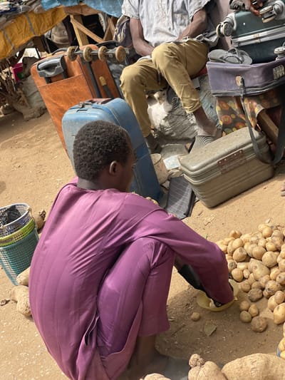 This image depicts an outdoor market scene in Zaria, Nigeria. In the foreground, a person with dark, short curly hair is squatting, viewed from behind, wearing a long-sleeved purple garment with subtle vertical stripes. This individual is positioned next to a pile of light-skinned potatoes or similar tubers on the sandy, dirt ground and appears to be handling or sorting them into a small yellow bowl. To their left, stacked light blue and lime green plastic baskets are visible.
In the mid-ground, behind the squatting figure, several pieces of luggage are stacked. These include an upright orange-brown wheeled suitcase, a light blue suitcase, a dark purple suitcase with a grey flip-flop resting on it, and a partially visible dark green suitcase. A rectangular, olive-green container with a latch is on the ground to the right of the purple-clad person.
Further back, partially obscured, a second person is seated, wearing a light-colored, possibly striped shirt and khaki pants. Their bare feet and a dark slide sandal are visible. The background reveals a makeshift shelter structure composed of wooden poles and draped tarpaulins or fabrics in blue and orange hues. The ground is consistently a reddish-brown dirt.