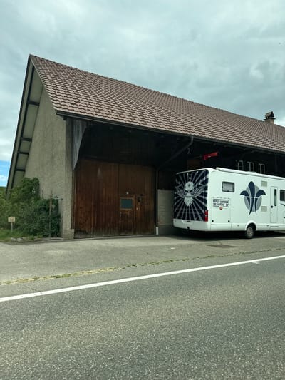 The image captures a white recreational vehicle (RV) parked alongside an asphalt road, next to a large building, under an overcast sky. The scene is set in Zurzach, Switzerland.

The RV is prominent, displaying extensive custom graphics. On its rear, a large blue and white design features an eagle-like bird with spread wings holding a skull or crest, against a sunburst background. Above this, a white rectangular plaque reads "HARLEY-DAVIDSON AARGAU," and a "CH" country sticker (indicating Switzerland) is visible below the eagle graphic. The side of the RV has an abstract blue and grey design.

To the left of the RV stands a substantial building, possibly a barn or agricultural structure, with a prominent brown, tiled gabled roof. The upper section of the building is constructed from dark wood, with small windows. Its lower part features large wooden garage doors, one of which has a small rectangular window. The base of the building is composed of a lighter-colored, textured wall, with some green bushes and foliage growing nearby.

An asphalt road occupies the right side of the frame, marked by a clear white solid line along its edge. Patches of grass and weeds are visible growing along the roadside. The sky above is uniformly grey and cloudy, indicating an overcast daytime. The RV appears to be parked or stored, with no human activity immediately apparent.