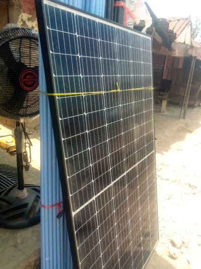 The scene features a solar panel leaning against a bundle of blue PVC pipes in what appears to be an outdoor location. A black stand fan with an orange logo sits to the left of the pipes. The background includes a partially visible building with a corrugated roof and sandy ground. The image was likely taken outdoors under natural lighting conditions in Minna, Nigeria. There are no people in the image. The solar panel is dark with a grid-like pattern. The pipes are bundled together with red plastic tape. A thin yellow string binds the solar panel to the pipes. The fan is of the oscillating type. The general environment is typical of a commercial or residential area with construction materials.