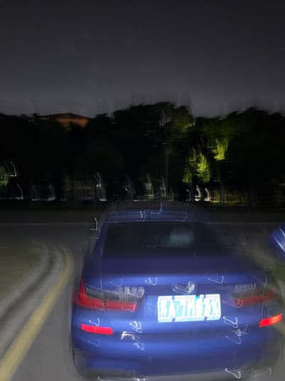 The image depicts a blurred scene at night, likely taken with a moving camera. A blue vehicle is partially visible on the right side of the frame, its taillights illuminated. To the left, there's a dark area suggesting trees or vegetation. There's also a sign on the vehicle, but the text is illegible due to the blur. The location is believed to be in the city of Jiangning District, China. Overall, it's a night scene with a focus on the vehicle and blurred surroundings.