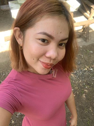 The image is a close-up, selfie-style shot of a young woman with medium-length auburn hair, taken outdoors. She is wearing a pink, ribbed t-shirt. She has light brown eyes, defined eyebrows, and a small mole on her right cheek. She is smiling, and her lips are colored with a reddish-brown lipstick. She is also wearing a square-shaped gold earring in her left ear. A clear, rectangular hair clip is attached to her t-shirt near her collarbone. The background appears to be a rural or outdoor setting, with dirt and gravel on the ground and some wooden structures visible behind her. The lighting suggests it is daytime, with sunlight casting shadows. The location is identified as Kiblawan, Philippines.
