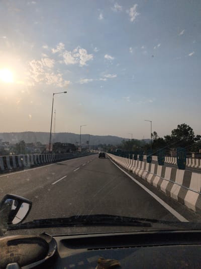 The image depicts a view from a vehicle traveling on a highway overpass in Samba, India. The sun is low in the sky, casting a warm, golden light and creating a haze. The highway is divided into multiple lanes, with concrete barriers adorned with black and white stripes along the sides. Streetlights are visible at intervals along the overpass. In the distance, hills form the horizon, and signs of urban development, including buildings and electrical poles, can be seen nestled at the base of the hills. Several cars are on the highway, indicating traffic flow. The interior of the vehicle is partially visible in the foreground, including the dashboard, a side mirror, and parts of the windshield. The overall impression is one of a serene, late afternoon journey on a highway.