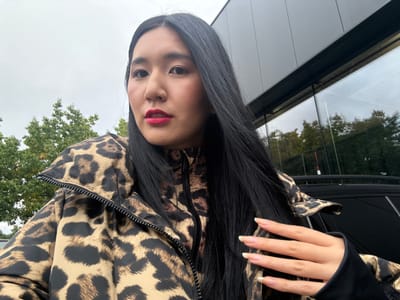 A female subject, appearing East Asian, with long, straight, dark hair and red lipstick, is depicted from the chest up in a selfie-style photograph taken in Herentals, Belgium. She is wearing a leopard print puffer jacket over a matching high-collared, zippered top. Her left hand is raised in the foreground, with fingers extended and natural-colored nails. The background on the left consists of green trees under an overcast sky. On the right, a modern building with dark grey paneling and large reflective glass windows is visible, reflecting trees and the sky. A dark-colored vehicle, possibly an SUV with a roof rack, is partially visible in the lower right behind the subject.