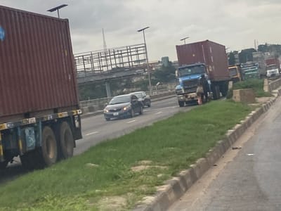 The image depicts a traffic scene on a highway, likely in Ikeja, Nigeria, with a cloudy sky overhead. In the foreground, a large red shipping container truck is partially visible on the left, with its wheels and lower chassis showing. To the right, a blue truck with a red shipping container is stopped, and a person in dark clothing carrying a light-colored bag is walking in front of it. Several other trucks, including a yellow truck, green and white trucks, and a white truck, are also part of the traffic jam, lined up on the right side of the road. A dark gray sedan is driving in the lane to the left of the blue truck. The road is bordered by a grassy embankment and a concrete curb. In the background, overhead bridges and structures can be seen, suggesting an urban or semi-urban environment. The overall impression is one of congestion and slow-moving traffic on a cloudy day.