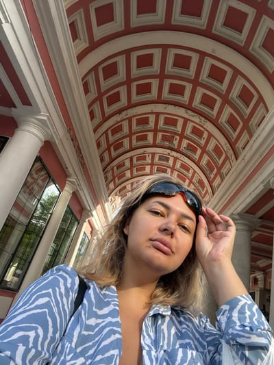 The image shows a woman taking a selfie outdoors. She is wearing a blue and white striped shirt and sunglasses perched on her forehead. Behind her, a red and white vaulted ceiling with square insets is visible, supported by large white columns and windows. This architectural style suggests a building in the city of Khabarovsk, Russia, possibly a public building or a historic site. The woman's hand is resting on her forehead, and she is looking towards the camera. The lighting indicates it is daytime, with sunlight casting shadows.