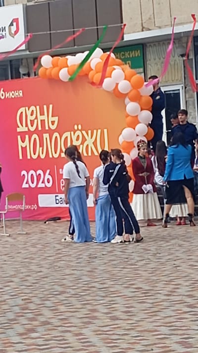 This media file captures an outdoor scene in Baksan, Russia, likely during a daytime event. The focal point is a large, vibrant banner that reads "ДЕНЬ МОЛОДЕЖИ 2026" in bold, yellow lettering against an orange and pink gradient background. This translates to "YOUTH DAY 2026," indicating a celebration of youth.

Several people are visible in the foreground, mostly young women and some men, dressed in a mix of casual and traditional attire. Some women are wearing long, flowing blue skirts and white tops, while others are in dark tracksuits. One woman is dressed in a striking red and gold traditional outfit, including a crown-like headpiece, suggesting a cultural performance or representation. Men in what appear to be uniforms, possibly security or official personnel, are also present, standing behind the banner.

The scene is decorated with a festive arch of orange and white balloons and colorful ribbons strung across the top. A paved ground, composed of interlocking bricks in shades of brown and beige, covers the area where the people are standing. In the background, a building with windows and signage can be seen.

The overall atmosphere appears to be one of celebration and gathering, likely for the "Youth Day" event. The lighting suggests it is daytime, and no specific weather conditions are discernible. Visible text includes the main banner, a smaller sign with the website "МОЛОДЕЖЬ.РФ", and partial text on the building in the background, one segment of which appears to be "НСКИЙ". The presence of traditional clothing alongside modern attire suggests a blend of cultural heritage and contemporary celebration.