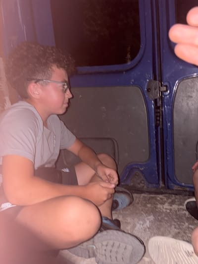 The image shows a close-up, low-angle view of a group of people in what appears to be an outdoor setting, possibly a park or recreational area in Kukorevo, Bulgaria. In the foreground, a young person with curly hair and glasses is lying down, looking upwards. They are wearing a grey t-shirt and black shorts. Another person's arm is visible across the image, and a hand is resting on the leg of the person lying down.

In the background, there are blue structures, possibly part of a playground or seating area, with dark, textured surfaces. A person's legs and feet are visible on the right side of the image, wearing grey athletic shoes. Another person's feet are visible in the upper left quadrant, wearing light-colored athletic shoes. The ground appears to be rough and textured, suggesting a natural or unpaved surface.

The lighting suggests it might be evening or nighttime, with artificial light possibly illuminating the scene. The overall mood seems relaxed and casual, possibly indicating a social gathering or downtime. No visible text is present in the image.