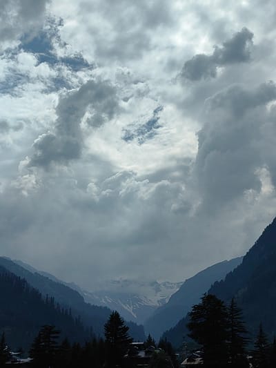 This media file depicts a dramatic mountain landscape under a tumultuous sky. The scene is dominated by layers of silhouetted pine trees and steep, dark mountain slopes that recede into the distance. In the far background, snow-capped peaks are visible, peeking through breaks in the clouds. The sky is filled with heavy, grey clouds, with some patches of lighter sky suggesting breaks in the overcast conditions. The lighting is dim, indicating it could be late afternoon or an overcast day. The overall mood is somber and dramatic, reflecting the powerful natural elements. No people or specific objects are discernible, making it a landscape-focused image. The location is suggested to be Manali, India, a region known for its Himalayan mountain scenery.