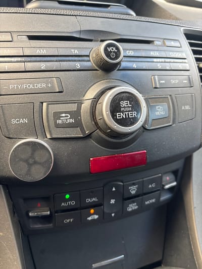 The image shows the interior of a car, specifically the center console and dashboard area. The focus is on the car's audio system and climate controls. The buttons and knobs of the stereo system are clearly visible, including controls for volume, CD, AUX, clock, tuning, and track selection. There are also buttons for menu, scan, return, and folder navigation. Below the stereo controls, there are buttons for climate control, including fan speed, mode selection, A/C, and dual climate zones, with indicators for "AUTO" and "OFF." Some of these climate control buttons have illuminated indicators. The dashboard is made of dark plastic and shows signs of dust accumulation. There is also a red hazard light button visible. The overall environment suggests the inside of a car that is parked. There are no people visible in the image, and no indication of the city of Lod, Israel.