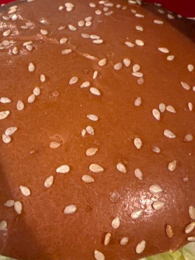 The image is a close-up, overhead shot of a sesame seed bun. The bun is a light brown color and is covered in numerous small, white sesame seeds. The texture of the bun appears slightly shiny and smooth. The image is tightly cropped, showing only the surface of the bun. There is no discernible context of location, including Rustavi, Georgia, within the visual information provided.