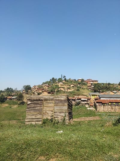 The image presents an overview of a hillside community in Kitsombiro, Democratic Republic of the Congo, under a clear blue sky with a faint crescent moon visible in the upper right. In the foreground, a rectangular structure constructed from horizontally stacked, weathered wooden logs stands on a patch of short green grass; a vertical wooden plank door is centered on its front face. The ground around this structure consists of dry earth and sparse vegetation, transitioning to thicker, taller grasses and uncultivated land. In the midground, the terrain begins to slope upwards, revealing scattered single-story buildings with various corrugated metal roofs, some in metallic silver, brown, or distinct yellow and green sections, partially enclosed by fences made of vertical wooden poles. The background is dominated by a densely populated, steep hill. Numerous small, simple dwellings with earthen-colored walls and reddish-brown roofs are clustered across the hillside, interspersed with green trees and broad-leafed plants, indicating a verdant environment. No individuals, animals, or specific actions are discernible within the frame.