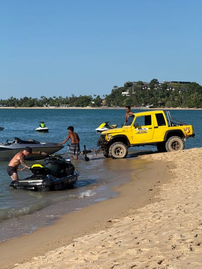 In Baan Mae Nam, Thailand, a bright yellow off-road vehicle is parked on a sandy beach next to a body of water. The vehicle has a roll cage and a spare tire mounted on the back. The driver's side door is open, and a man is visible inside the vehicle. Several jet skis are present on the water and near the shore. Three men are interacting with the jet skis. One man is standing in the water near a jet ski, and another man is on the shore, looking at a jet ski. A third man, who is tattooed, is sitting on a jet ski. The scene appears to be set during the daytime, with clear blue skies. The beach is lined with lush green trees and buildings in the background, suggesting a tropical location. The overall mood seems to be relaxed and recreational, with people enjoying activities on the water. A sticker on the vehicle reads "OFF ROAD." Another visible text is "Valvoline" on the side of the yellow vehicle.