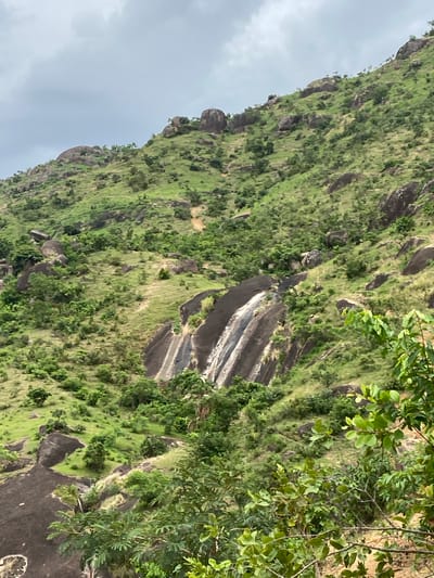 The image depicts a steep, rocky hillside covered in lush green vegetation in Ganawuri, Nigeria. The landscape is dotted with large boulders and patches of grass and shrubs. A prominent rock formation with distinct, light-colored striations runs vertically down the slope. The sky appears overcast with a thick cloud cover visible on the left side of the image. Branches and leaves from a tree or bush are visible in the foreground on the right.