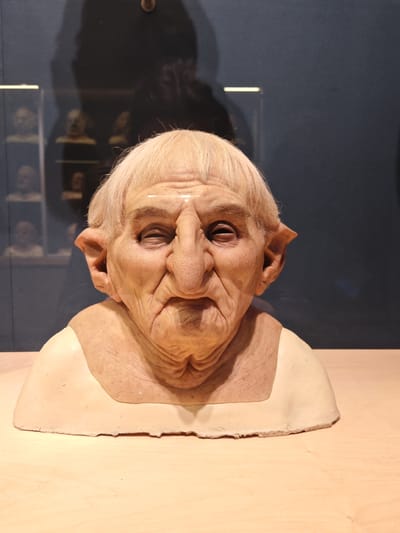 A bust of an elderly person with a pronounced nose and pointed ears is displayed on a light-colored wooden surface. The bust has pale skin and short, white hair. The backdrop is a dark blue wall, reflecting a shadowy figure and a display case filled with similar busts in the background. The image was taken indoors. The location is the city of Watford, United Kingdom.
