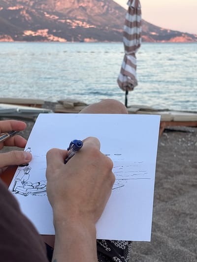 A person is drawing on a sheet of white paper with a blue pen, sitting on a beach. The drawing depicts two figures on a beach chair. The background shows the sea, a closed umbrella, and hills in the distance. The scene is set in Bečići, Montenegro. The lighting suggests it might be late afternoon or early evening. The person is wearing patterned shorts.