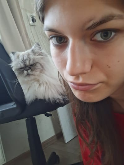 A woman is looking down into the camera with a slight smile. Below her is a cat, possibly a Persian or Himalayan breed, sitting in what appears to be a black office chair. The background includes a wall, possibly paneled or striped. The location is not possible to determine based on the available information.