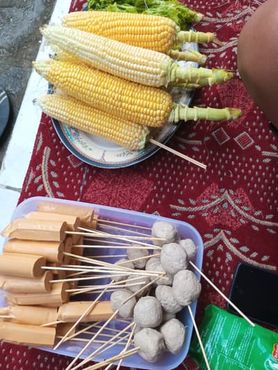 The scene in Ciherang, Indonesia, captures an arrangement of food items on a patterned red rug, adjacent to a white tiled floor. In the upper portion, a ceramic plate with a blue and white pattern holds five corn cobs; four are yellow, and one is a lighter pale yellow, all with trimmed green husks. Green leafy vegetables are visible behind the plate. Below the corn, a clear plastic rectangular container is filled with skewered items: approximately 15 light brown cylindrical sausages on wooden sticks are arranged on the left, and roughly 20 greyish spherical meatballs on wooden sticks are positioned on the right. A dark rectangular object, possibly a smartphone, and a partially visible green package bearing "Del Monte" branding are present on the red rug to the right of the container. The upper right edge of the frame shows a segment of a human leg with dark skin. The environment appears to be an outdoor or semi-outdoor setting under natural light.