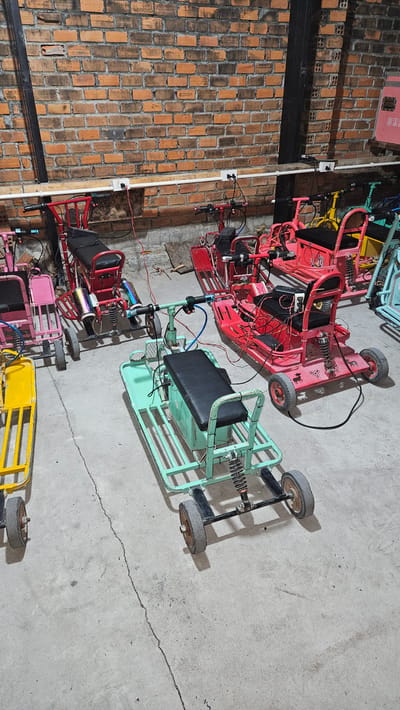 The image captures an indoor scene featuring multiple small, custom-built recreational vehicles, resembling go-karts or electric scooters, parked on a concrete floor. On the left, a textured brick wall with a dark horizontal beam spans the length of the image. White electrical conduits with visible outlets are mounted vertically on the brick wall, suggesting a utility or storage area.
There are at least five such vehicles, distinct in color. The most prominent one in the upper middle is red, featuring a metal frame, a black seat, exposed wiring, and rubber wheels. Below it, a light blue or mint green vehicle, also with a black seat and exposed components, appears to be connected to the red one by wires. Further down, parts of a pink and a yellow vehicle are visible. All vehicles exhibit a robust, somewhat industrial design with visible suspension springs, steering mechanisms, and numerous wires, suggesting they are electric-powered or perhaps undergoing maintenance/charging. Some vehicles show signs of use, with scuff marks on wheels and frames.
The concrete floor displays cracks, indicating wear. No people are present, and no specific activity is clearly depicted beyond the vehicles being parked. The lighting is even, suggesting an indoor setting or diffused light, so the time of day and weather are not discernible. No clear, legible text is visible anywhere in the image. The scene suggests a workshop, garage, or storage facility for these vehicles, located in Cam Đức, Vietnam.