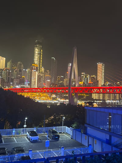 This is a nighttime, wide-angle shot of a city skyline with a prominent red bridge. The cityscape is illuminated with numerous lights from tall buildings, creating a vibrant and dynamic scene. The red bridge, with its intricate structure and illuminated cables, stands out against the dark sky and the bright city lights.
The image shows a modern urban environment, likely a downtown area or a well-developed district. Trees are visible in the foreground, suggesting a mix of urban development and green spaces. In the right foreground, a parking lot with several cars and charging stations is visible, indicating a commercial or residential area. The blue lighting on the building in the right foreground adds to the modern aesthetic.
The time of day is clearly nighttime, as evidenced by the dark sky and the extensive artificial lighting. The weather appears clear, with no visible precipitation or fog. The presence of Chinese characters on some of the buildings confirms the location as China.
There are no people visible in the image. The overall mood conveyed is one of urban energy and modernity, with the illuminated cityscape and bridge creating a visually striking spectacle. The combination of architectural elements and lighting design suggests a lively and prosperous urban center.