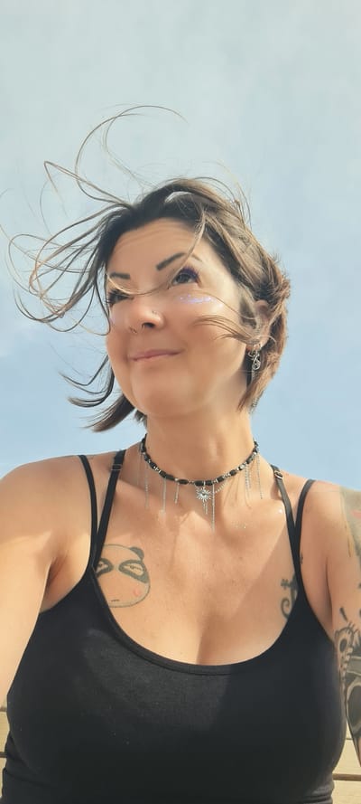 A low-angle, close-up shot captures a person against a bright, clear blue sky, likely outdoors in Nahariyya, Israel, during daytime. The individual is looking down towards the camera with a subtle, content smile.

The subject is a female-presenting individual with short, dark brown hair, some strands of which are softly blowing. She wears distinctive makeup, featuring purple eyeshadow and iridescent glitter flakes prominently on her left cheekbone and around her eye. Facial piercings include a small stud on her nose and a hoop through her septum.

She is dressed in a black tank top or bralette with thin straps. Adorning her neck is a unique black beaded necklace with multiple delicate silver chains and a star-shaped charm, with some chains cascading onto her upper chest. She also wears silver, chain-style earrings.

Numerous tattoos are visible on her arms, shoulders, and chest. A cartoon-like tattoo can be seen on her right chest/upper breast area, while her left arm and shoulder are extensively covered with intricate black and grey designs, including elements resembling skulls. Scattered glitter is also visible on her skin, suggesting a festive or artistic touch. No text is visible in the image.