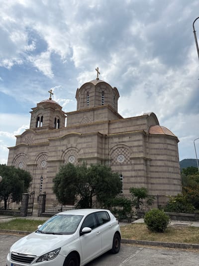 The image shows the exterior of a large stone church with striped patterns and domes. A white car is parked on the right side of the image, partially obscuring the lower portion of the church. The sky is cloudy, with patches of blue sky visible. Trees and greenery surround the church. The time of day appears to be daytime. There is no visible text in the image.