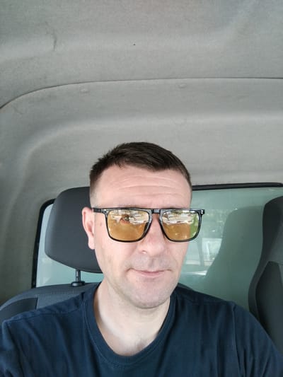 The image shows a close-up, selfie-style portrait of a man wearing sunglasses and a dark t-shirt, taken from inside a vehicle. The man has short, dark hair and is looking slightly down and to his left. The sunglasses have thick, dark frames and amber-tinted lenses which reflect a scene of trees, buildings, and a road. The background of the image shows the interior of a vehicle, with a headrest of a seat visible behind the man's head and the light-colored, textured ceiling of the vehicle above. A window with a greenish tint is visible to the right, and a glimpse of another seat can be seen. The lighting suggests it is daytime, and the reflections in the sunglasses indicate an outdoor environment, possibly a suburban or rural area. The overall mood is casual and everyday. The image was taken in Kitzingen, Germany. No visible text is present in the image.