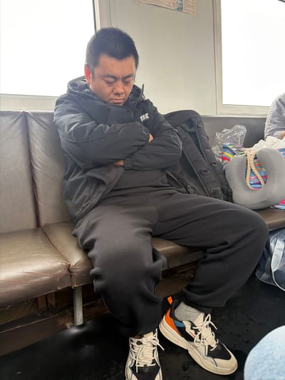 An overhead shot captures a man sleeping on a brown leatherette bench seat, likely within a mode of transport such as a train or bus, potentially in or around Nizhneleninskoe, Russia. The man is lying on his left side, facing towards the left of the frame, with his eyes closed and arms crossed over his chest. He is wearing a black puffer jacket, black pants, and grey socks. His feet, clad in white, black, blue, and orange athletic shoes, rest near the edge of the seat, with a dark, possibly wet, floor visible below.

Beside his legs and behind his head, a black backpack and a grey neck pillow with colorful striped straps are visible. Other bags, one partly covered in clear plastic and another light blue, are also present, suggesting travel. The seating appears worn, with visible cracks and creases in the leatherette. To the left of the man, a large, bright white window indicates daylight, possibly an overcast sky. A grey wall runs alongside the window, and a crumpled document or newspaper with indecipherable text is affixed to it. A metal pipe runs along the bottom of the seat structure, near the dark floor. The overall impression is one of rest during a journey.