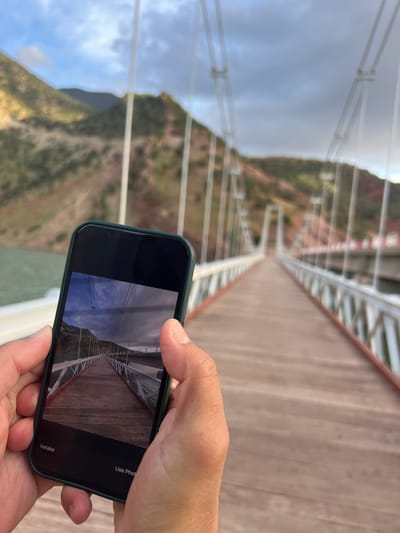 A person is holding a smartphone with a green case, displaying a picture of a long, wooden bridge. The person is standing on the same bridge, as can be seen in the background, which includes the bridge's wooden planks and the cable railings. The bridge is suspended over a mountainous landscape with a cloudy sky above. Based on the colors of the mountains the location is near the city of Tabaroucht, Morocco.