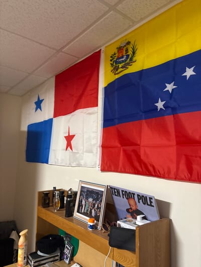 Two flags adorn a wall. The upper flag displays yellow, blue, and red stripes with white stars and a coat of arms. The flag below is white with red and blue corners and a red star on the upper side, plus a blue star on the lower. The flags are mounted next to a wooden desk. The desk contains various objects including framed pictures, cologne, and a book cover that says "TEN FOOT POLE." The image is rotated clockwise.