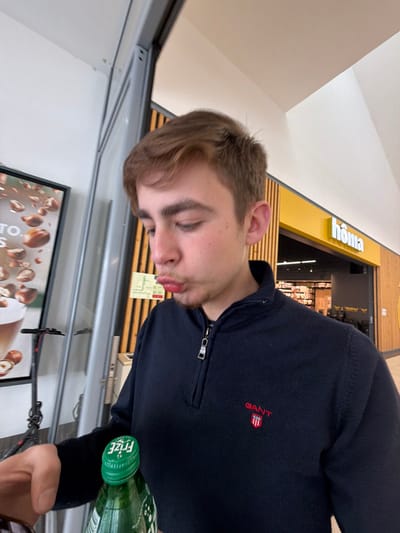 A young male, estimated to be in his late teens or early twenties, is depicted from the mid-chest upwards in an indoor commercial setting, likely a shopping mall in Vila Real, Portugal. He has short, light brown hair and a light complexion, presenting a pouting facial expression with puckered lips while looking downwards and to his right. He is wearing a navy blue quarter-zip sweater with a small red and white GANT shield logo on the left chest. His left hand is visible in the lower left foreground, holding a green plastic bottle with a matching green cap, clearly marked with the inverted white text "Frize."
In the background to the left, a portion of a large vertical display or poster is visible, advertising a beverage with scattered hazelnuts and partial text. Below this display, the handlebars and front wheel of a black and silver electric scooter are present. Directly behind the male subject, a wall features vertical light-toned wooden slats. To the right of the subject, a retail storefront is visible, identified by a prominent yellow sign displaying "hômə" in white lettering, with merchandise visible within the store's interior. The ceiling above is white and angled, indicative of modern commercial architecture, and the area is brightly lit by overhead lighting.